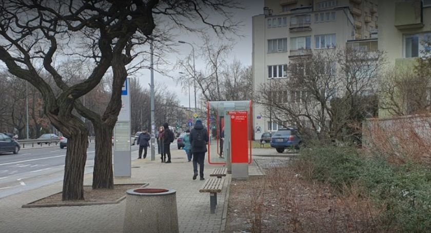 Przystanek autobusowy w Warszawie, na Mokotowie. Przechodzący chodnikiem ludzie, kosz na śmieci, w tle budynki.